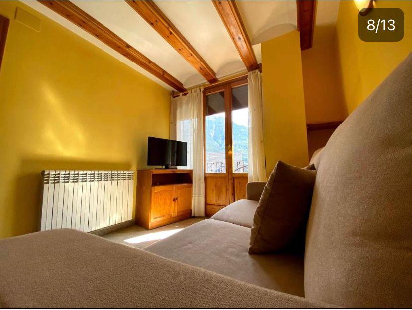 Sala de estar acogedora con sofá, televisor y ventana que ofrece vistas a las montañas. Pared amarilla y techos con vigas de madera, creando un ambiente cálido y luminoso. Radiador visible para calefacción.