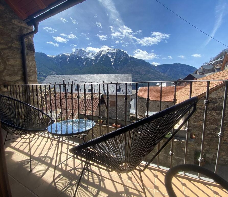Balcón con vistas a montañas y casas de piedra, equipado con sillas modernas y una mesa de vidrio, en un día soleado.