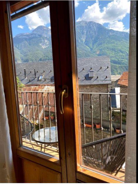 Vista desde una ventana de madera que muestra un balcón con una mesa y sillas, rodeado de casas de piedra y techos de tejas, con montañas verdes y un cielo azul de fondo.
