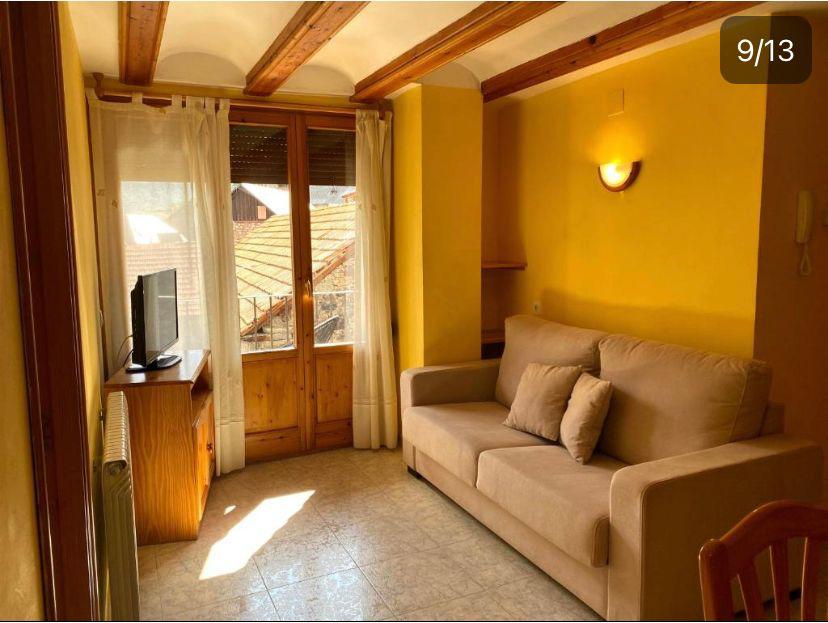 Sala de estar luminosa con paredes amarillas, sofá cómodo y mesa de madera, equipada con televisión y ventanas que ofrecen vistas al exterior. Ideal para relajarse y disfrutar de la luz natural.