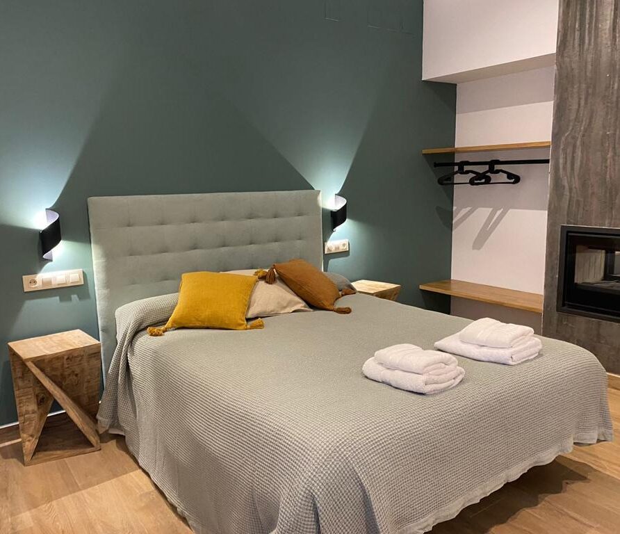 Habitación moderna con cama doble, decorada en tonos verdes y beige, con mesitas de noche de madera, toallas blancas y una chimenea elegante. Ideal para una estancia acogedora y relajante.