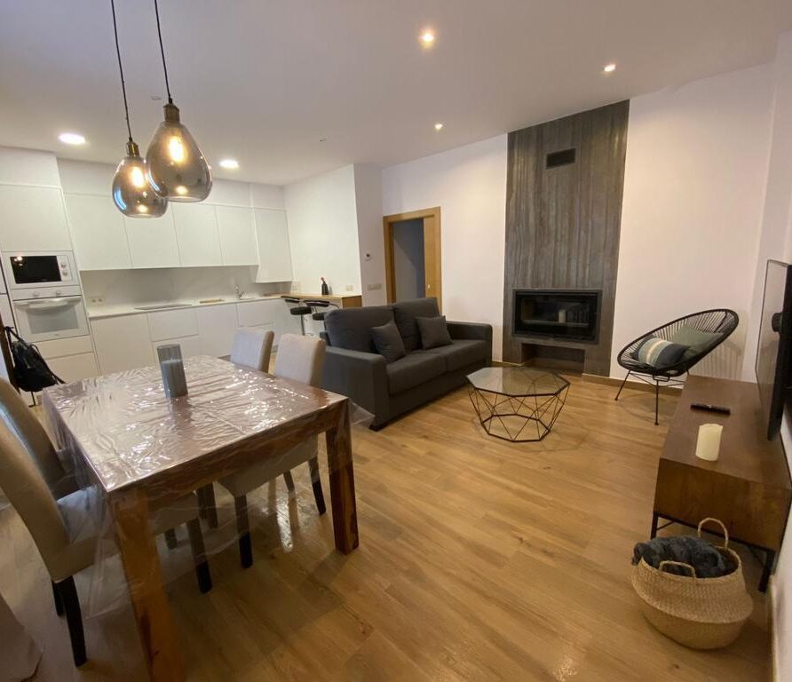 Sala de estar moderna con cocina abierta, mesa de comedor de madera, sofá gris, chimenea y decoración minimalista. Iluminación cálida y suelo de madera, creando un ambiente acogedor.