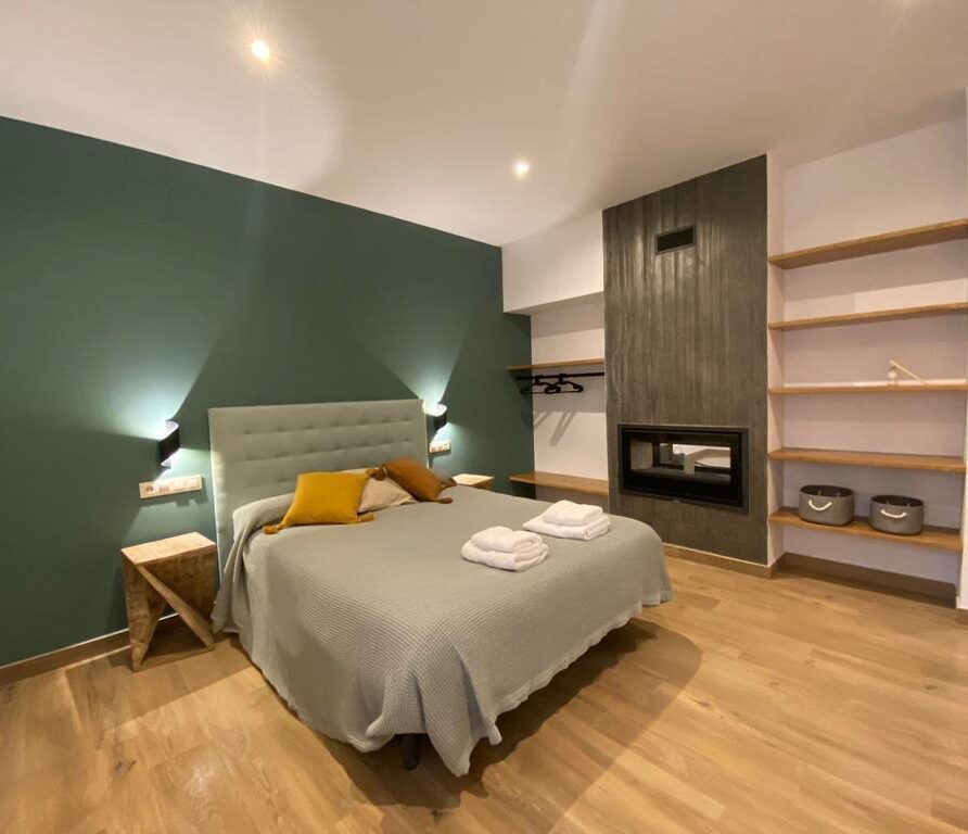 Habitación moderna con cama doble, paredes verdes y decoraciones minimalistas. Incluye chimenea, mesas de noche y estanterías de madera. Ideal para una escapada relajante.