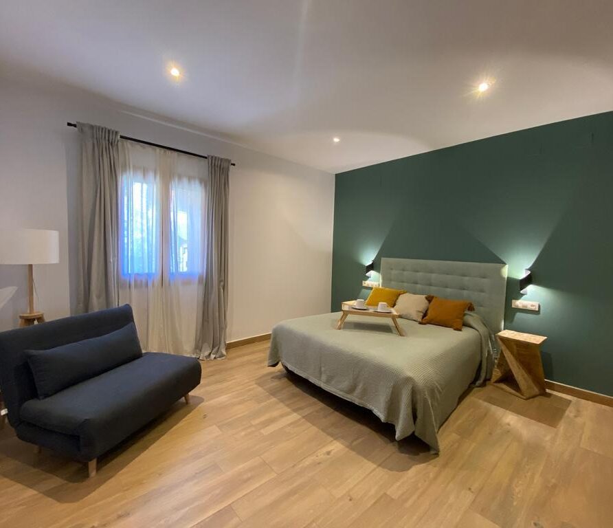 Habitación moderna con cama doble, decoración en tonos neutros, sofá negro y ventana con cortinas. Ideal para descansar y disfrutar de un ambiente acogedor.