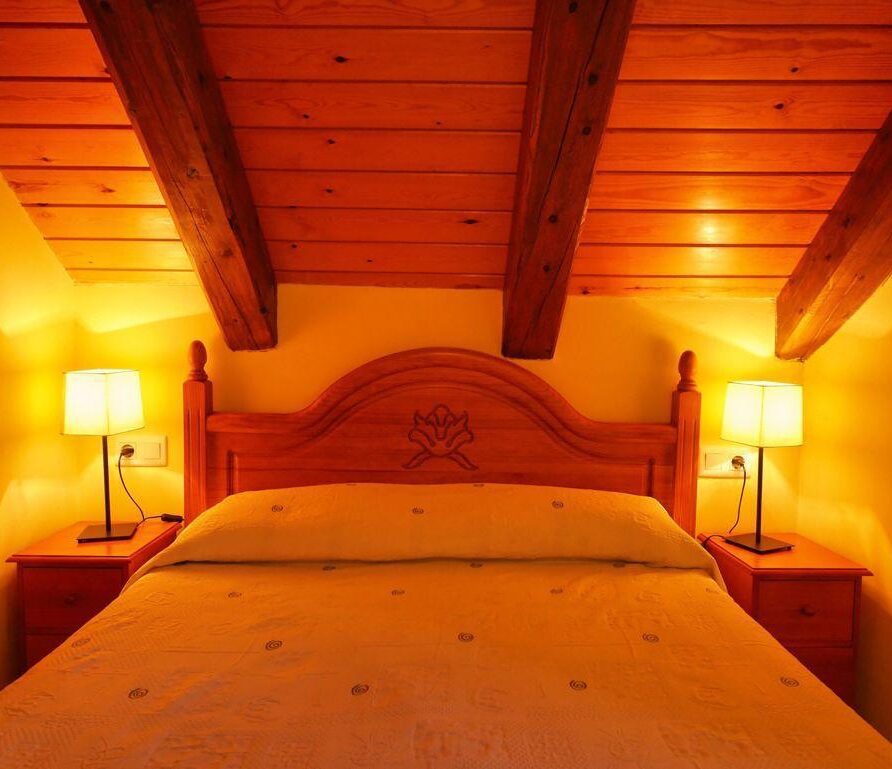Habitación acogedora con cama doble, cabecera de madera tallada y lámparas de noche, decorada en tonos cálidos y con techos de madera expuesta. Ideal para un descanso confortable.