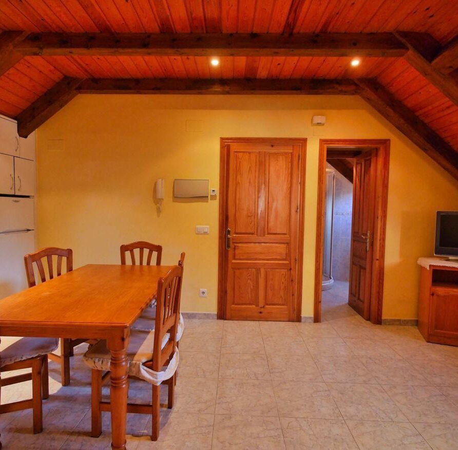Interior de un apartamento acogedor con techo de madera, mesa de comedor de madera y sillas, cocina equipada y una puerta que lleva a un baño.