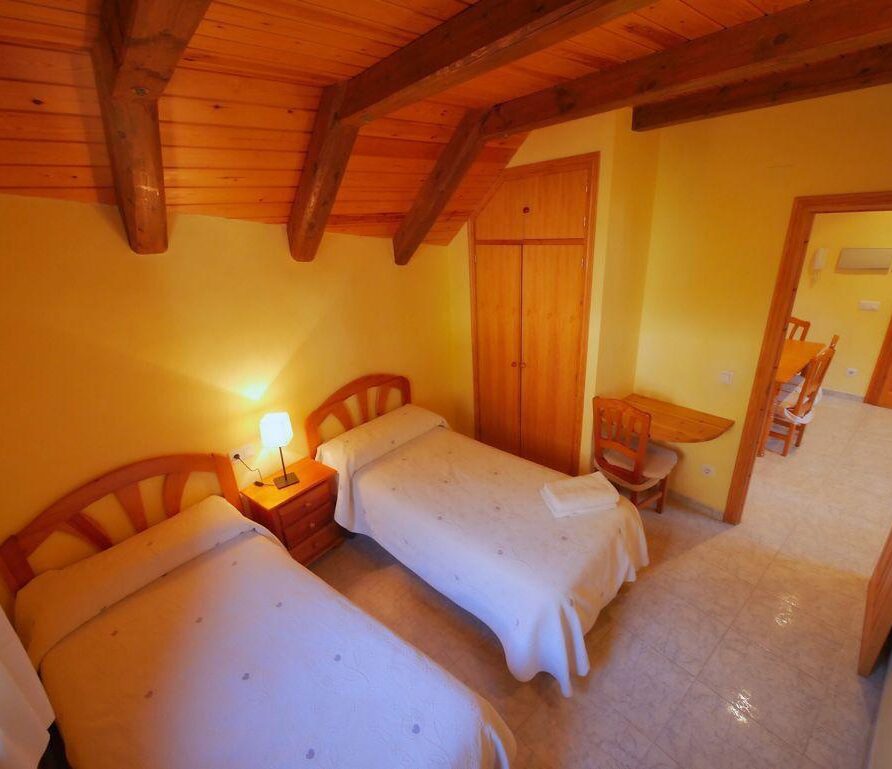 Habitación acogedora con dos camas individuales, decorada en tonos amarillos y madera, iluminada por una lámpara de mesa. Incluye un armario y un pequeño escritorio con sillas, ideal para descansar o trabajar.