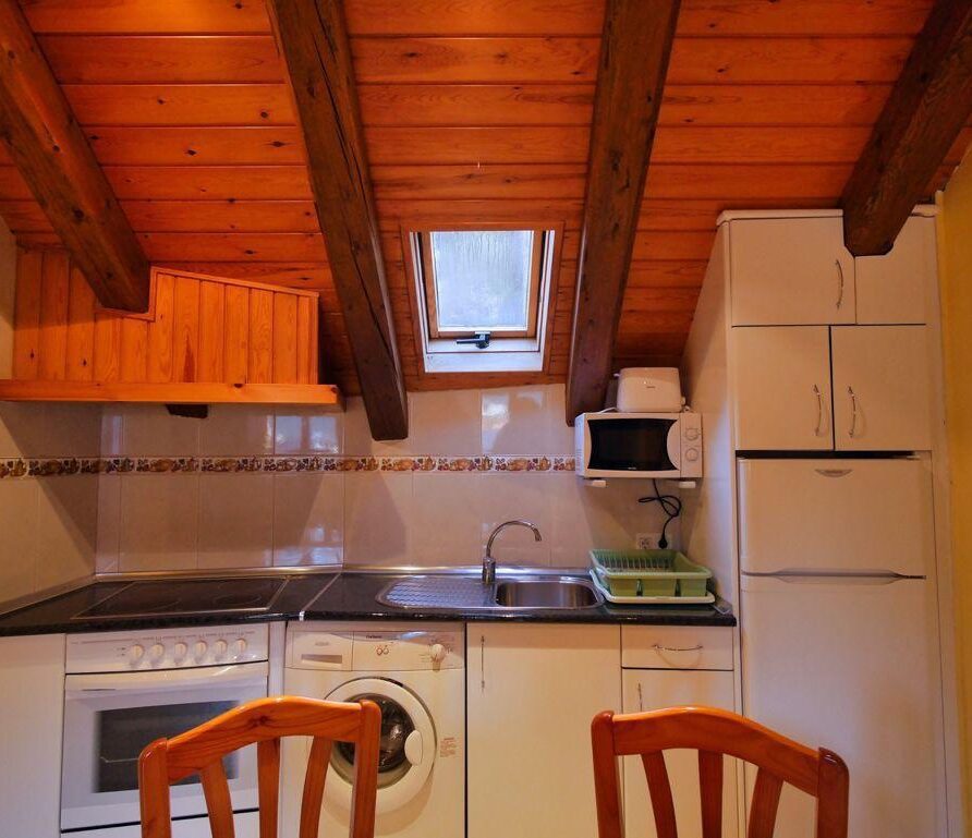 Cocina moderna con techo de madera, equipada con electrodomésticos como horno, lavadora y microondas, además de un fregadero y espacio de almacenamiento. La iluminación natural entra por una ventana en el tejado.