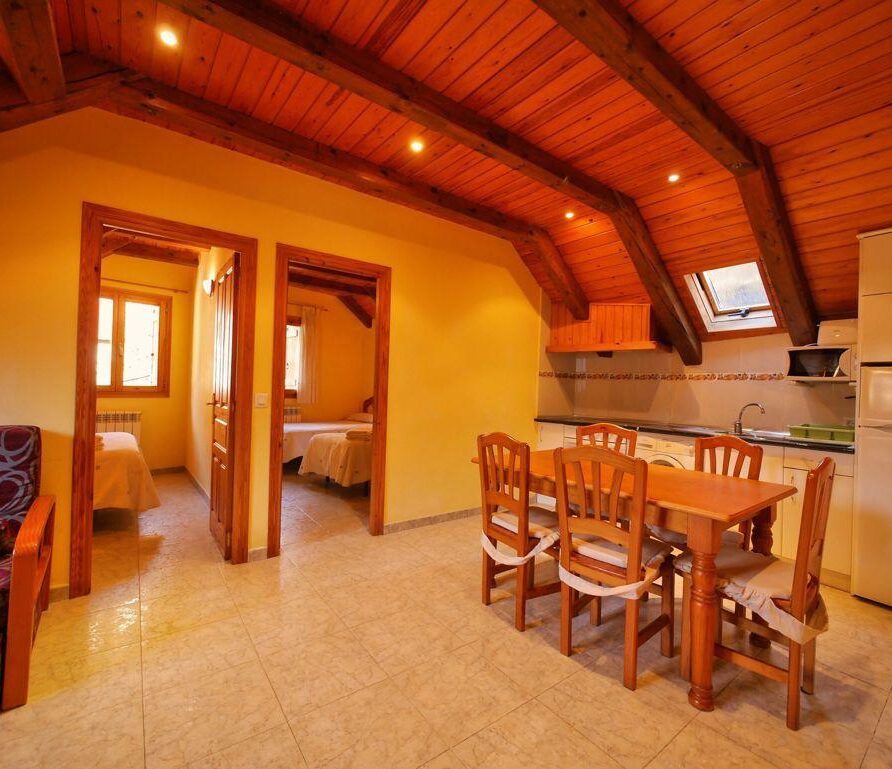Interior de un apartamento acogedor con techos de madera, que incluye una sala de estar con sofá, un área de comedor con mesa y sillas, y una cocina equipada. Se observan dos habitaciones con camas individuales al fondo, iluminadas por ventanas. Ideal para estancias familiares o grupales.