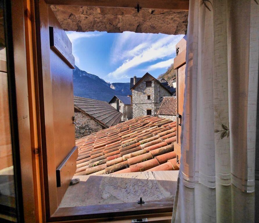 Vista desde una ventana de madera que muestra techos de tejas rojas y casas de piedra en un pueblo montañés, con un cielo azul y montañas al fondo.