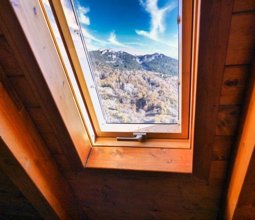 Ventana de techo que muestra un paisaje montañoso con árboles y cielo azul, destacando la estructura de madera del interior.