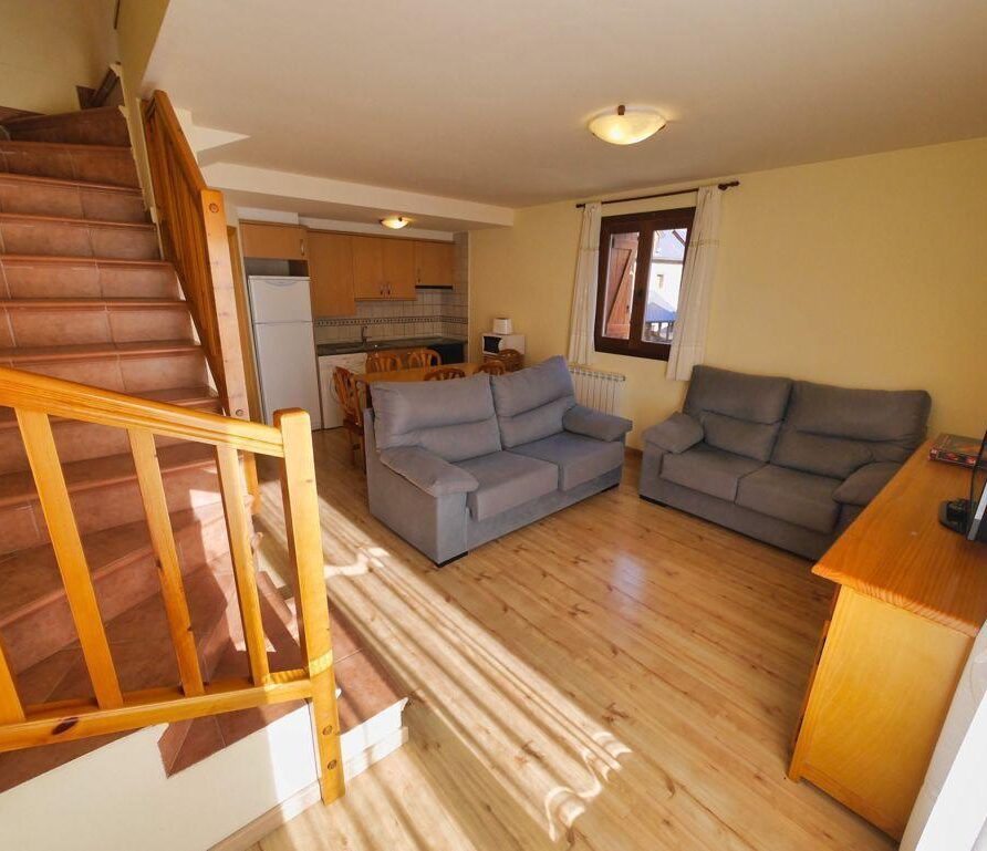 Sala de estar acogedora con escaleras de madera, dos sofás grises, una mesa de comedor y cocina equipada, iluminada por luz natural. Ideal para descansar y disfrutar en familia.