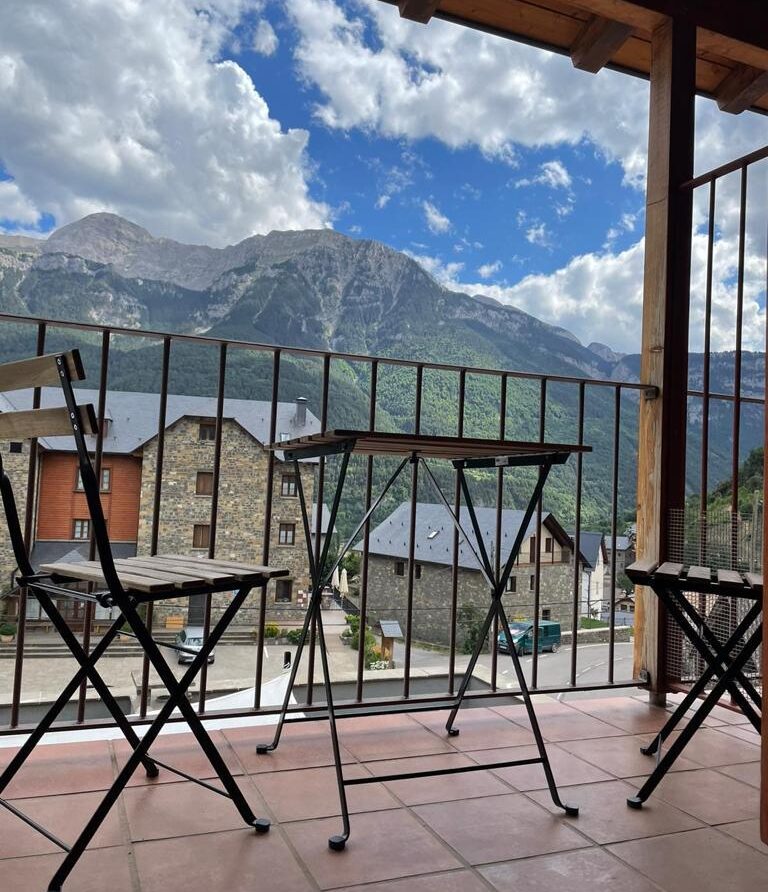 Vista panorámica desde un balcón con sillas y mesa de metal, mostrando montañas imponentes y un pueblo pintoresco en el fondo, bajo un cielo nublado. Ideal para escapadas en la naturaleza.