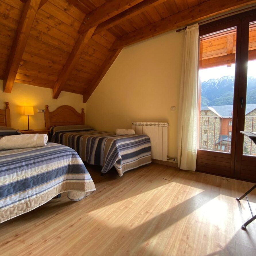 Habitación acogedora con techos de madera, dos camas individuales con sábanas a rayas, luz cálida, y ventana que ofrece vistas a montañas. Ideal para una escapada tranquila.