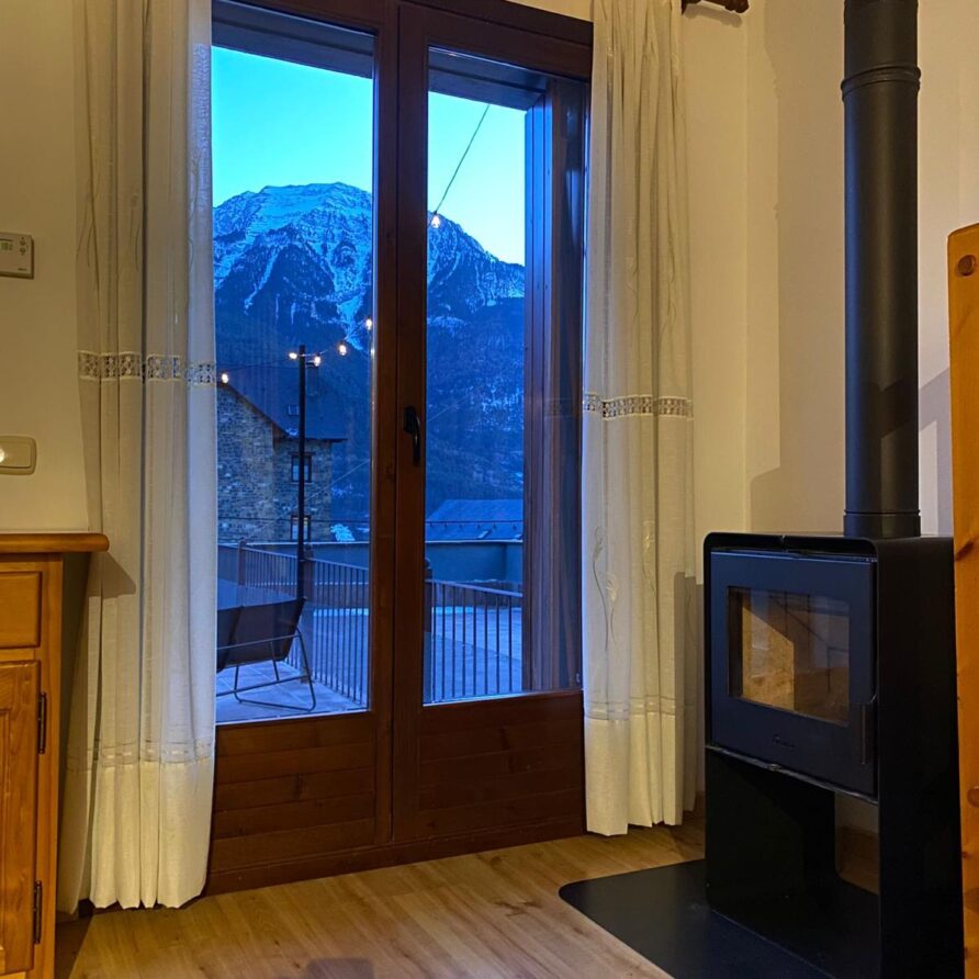 Interior acogedor con ventana que muestra montañas nevadas al atardecer, estufa de leña a un lado y muebles de madera. Ideal para disfrutar de la naturaleza y el confort en un ambiente rural.