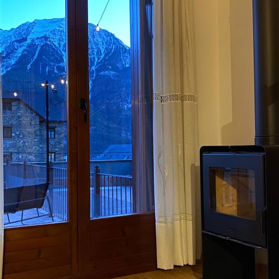 Vista interior de una cabaña con una estufa moderna, mostrando una ventana grande que da a un paisaje montañoso nevado al atardecer. La decoración incluye cortinas blancas y un suelo de madera, creando un ambiente acogedor y cálido.