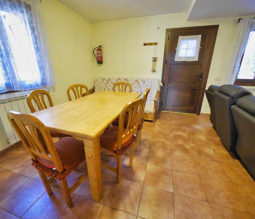 Sala de estar acogedora con mesa de madera y sillas, sofá de cuero, ventana con cortinas y puerta de madera, ideal para reuniones familiares.