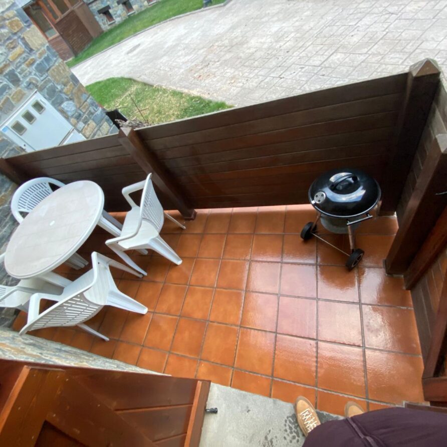 Terraza con mesa redonda y cuatro sillas blancas, ideal para comidas al aire libre, junto a una parrilla negra. Fondo de piedra y césped, creando un ambiente acogedor.