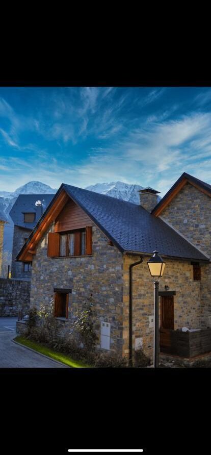 Casa de piedra con tejado a dos aguas, ubicada en un entorno montañoso. El cielo azul y despejado resalta la arquitectura de la vivienda, que cuenta con ventanas de madera y un farol en el jardín. Ideal para escapadas en la naturaleza.