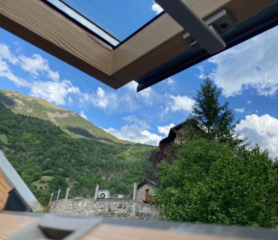 Vista panorámica desde una ventana de madera, mostrando montañas verdes y un cielo azul con nubes, con una casa rústica en el fondo. Ideal para ilustrar paisajes naturales y arquitectura en entornos montañosos.
