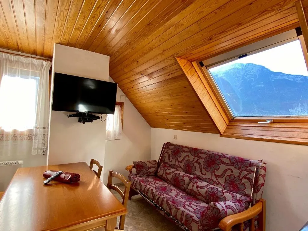 Interior de una habitación acogedora con techo de madera, sofá cama con estampado floral, mesa de madera y televisor montado en la pared. Ventana con vista a las montañas y luz natural que ilumina el espacio.