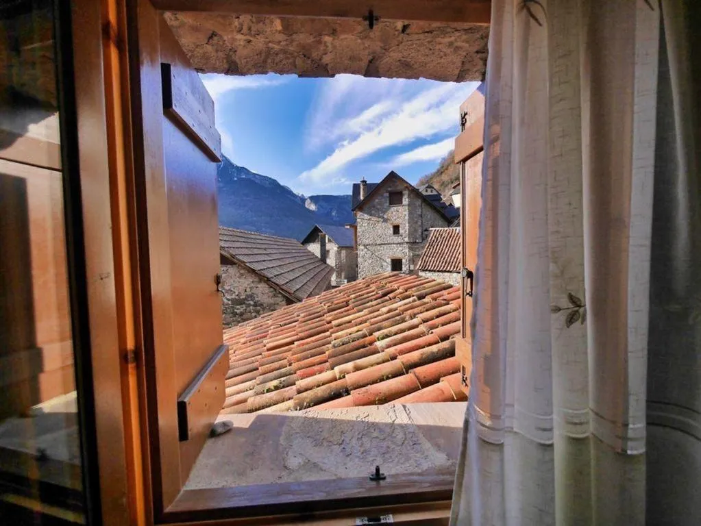 Ventana abierta con vistas a un paisaje montañoso y techos de tejas en un pueblo pintoresco, iluminado por la luz natural. La escena muestra la tranquilidad de la arquitectura tradicional en un entorno rural.